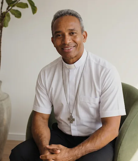 Padre Vanilson Silva, de camisa clerical branca e crucifixo, sentado e sorrindo